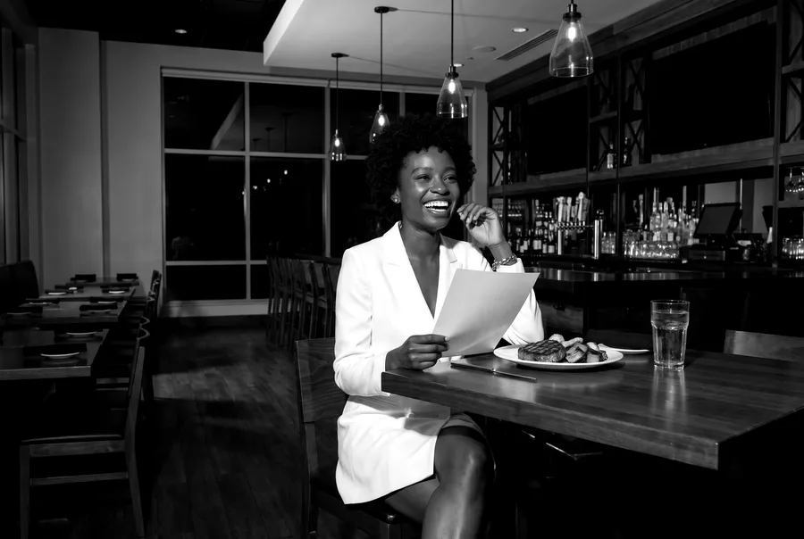 Black-and-white generated image: the guide's character laughs at a table in a bar and grill, a menu in her hand and a plate of steak and vegetables in front of her.